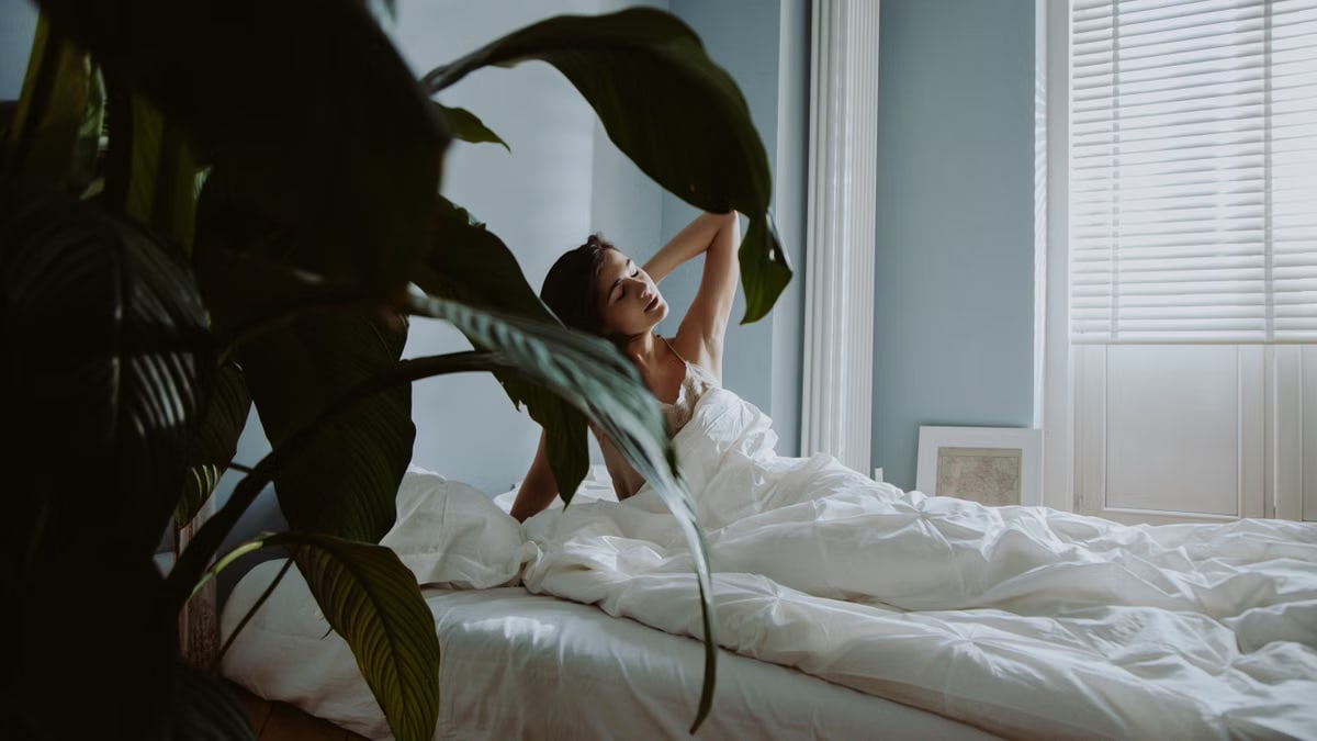A woman waking up in a cozy bedroom with natural light and lush greenery.